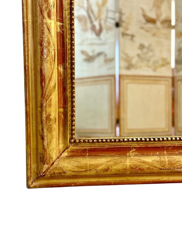 This exquisite French Louis Philippe mirror dates from the mid-19th century, around 1850, and exemplifies the elegant simplicity characteristic of the Louis Philippe period. The frame features rounded upper corners and a rectangular silhouette, enhanced by its rich gold leaf finish over red bole. Subtle floral engravings are etched into the gesso, adding a touch of refinement without overpowering the frame’s classic lines. The mirror’s warm patina is accentuated by signs of age and wear, including a few small losses and touch-ups to the gilding, particularly visible on the edges and corners, which have been sympathetically restored in the past. These time-worn details contribute to the mirror’s authentic antique charm and are consistent with its age and provenance. The glass is original, with light foxing and a soft, reflective surface typical of 19th-century mercury-backed mirrors. It is surrounded by a delicate beaded border that adds further depth and definition to the frame. With its understated elegance and generous size, this mirror would suit both classic and contemporary interiors. It can be placed above a fireplace, console table, or used as a floor mirror for added dramatic effect. A timeless piece that brings a touch of French sophistication to any space.