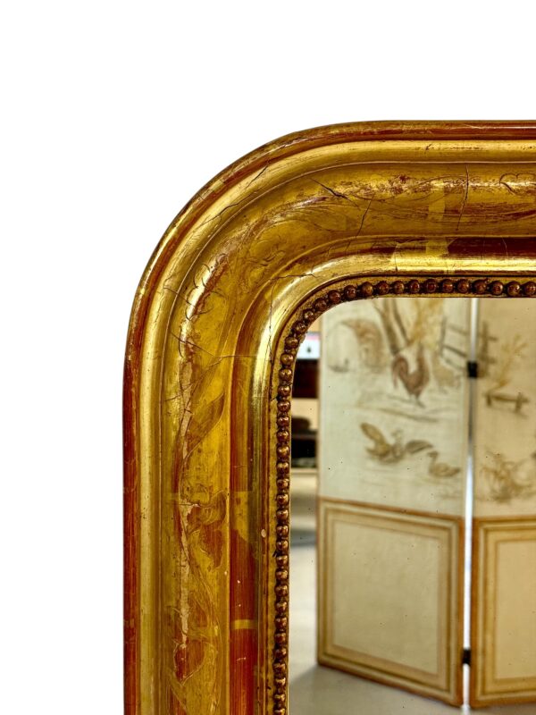 This exquisite French Louis Philippe mirror dates from the mid-19th century, around 1850, and exemplifies the elegant simplicity characteristic of the Louis Philippe period. The frame features rounded upper corners and a rectangular silhouette, enhanced by its rich gold leaf finish over red bole. Subtle floral engravings are etched into the gesso, adding a touch of refinement without overpowering the frame’s classic lines. The mirror’s warm patina is accentuated by signs of age and wear, including a few small losses and touch-ups to the gilding, particularly visible on the edges and corners, which have been sympathetically restored in the past. These time-worn details contribute to the mirror’s authentic antique charm and are consistent with its age and provenance. The glass is original, with light foxing and a soft, reflective surface typical of 19th-century mercury-backed mirrors. It is surrounded by a delicate beaded border that adds further depth and definition to the frame. With its understated elegance and generous size, this mirror would suit both classic and contemporary interiors. It can be placed above a fireplace, console table, or used as a floor mirror for added dramatic effect. A timeless piece that brings a touch of French sophistication to any space.