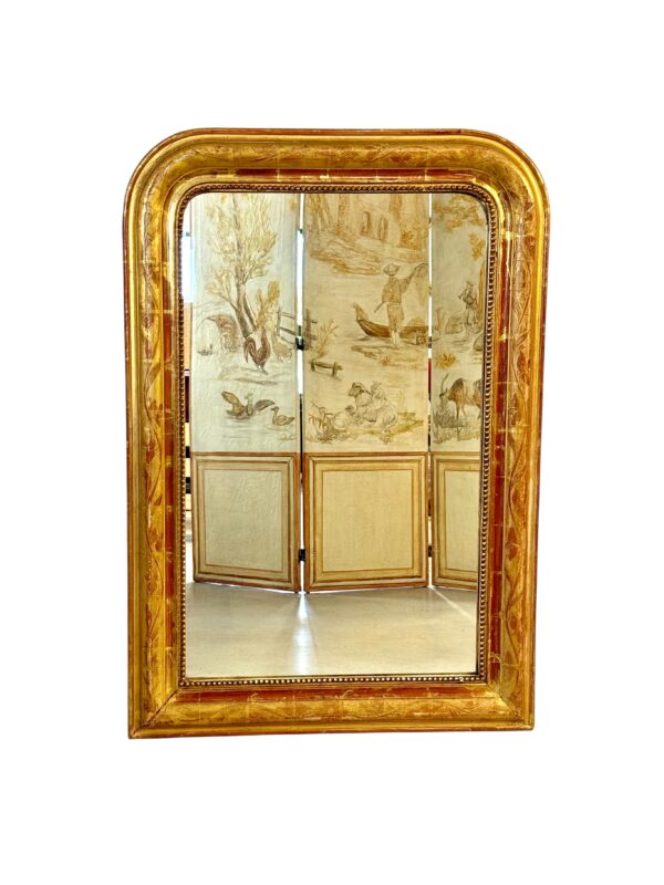 This exquisite French Louis Philippe mirror dates from the mid-19th century, around 1850, and exemplifies the elegant simplicity characteristic of the Louis Philippe period. The frame features rounded upper corners and a rectangular silhouette, enhanced by its rich gold leaf finish over red bole. Subtle floral engravings are etched into the gesso, adding a touch of refinement without overpowering the frame’s classic lines. The mirror’s warm patina is accentuated by signs of age and wear, including a few small losses and touch-ups to the gilding, particularly visible on the edges and corners, which have been sympathetically restored in the past. These time-worn details contribute to the mirror’s authentic antique charm and are consistent with its age and provenance. The glass is original, with light foxing and a soft, reflective surface typical of 19th-century mercury-backed mirrors. It is surrounded by a delicate beaded border that adds further depth and definition to the frame. With its understated elegance and generous size, this mirror would suit both classic and contemporary interiors. It can be placed above a fireplace, console table, or used as a floor mirror for added dramatic effect. A timeless piece that brings a touch of French sophistication to any space.