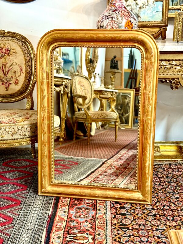 This exquisite French Louis Philippe mirror dates from the mid-19th century, around 1850, and exemplifies the elegant simplicity characteristic of the Louis Philippe period. The frame features rounded upper corners and a rectangular silhouette, enhanced by its rich gold leaf finish over red bole. Subtle floral engravings are etched into the gesso, adding a touch of refinement without overpowering the frame’s classic lines. The mirror’s warm patina is accentuated by signs of age and wear, including a few small losses and touch-ups to the gilding, particularly visible on the edges and corners, which have been sympathetically restored in the past. These time-worn details contribute to the mirror’s authentic antique charm and are consistent with its age and provenance. The glass is original, with light foxing and a soft, reflective surface typical of 19th-century mercury-backed mirrors. It is surrounded by a delicate beaded border that adds further depth and definition to the frame. With its understated elegance and generous size, this mirror would suit both classic and contemporary interiors. It can be placed above a fireplace, console table, or used as a floor mirror for added dramatic effect. A timeless piece that brings a touch of French sophistication to any space.