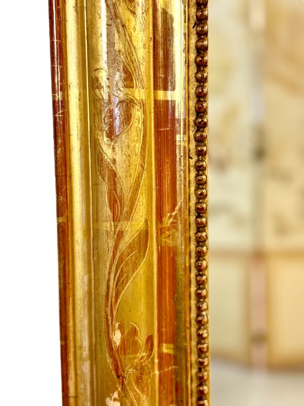 This exquisite French Louis Philippe mirror dates from the mid-19th century, around 1850, and exemplifies the elegant simplicity characteristic of the Louis Philippe period. The frame features rounded upper corners and a rectangular silhouette, enhanced by its rich gold leaf finish over red bole. Subtle floral engravings are etched into the gesso, adding a touch of refinement without overpowering the frame’s classic lines. The mirror’s warm patina is accentuated by signs of age and wear, including a few small losses and touch-ups to the gilding, particularly visible on the edges and corners, which have been sympathetically restored in the past. These time-worn details contribute to the mirror’s authentic antique charm and are consistent with its age and provenance. The glass is original, with light foxing and a soft, reflective surface typical of 19th-century mercury-backed mirrors. It is surrounded by a delicate beaded border that adds further depth and definition to the frame. With its understated elegance and generous size, this mirror would suit both classic and contemporary interiors. It can be placed above a fireplace, console table, or used as a floor mirror for added dramatic effect. A timeless piece that brings a touch of French sophistication to any space.